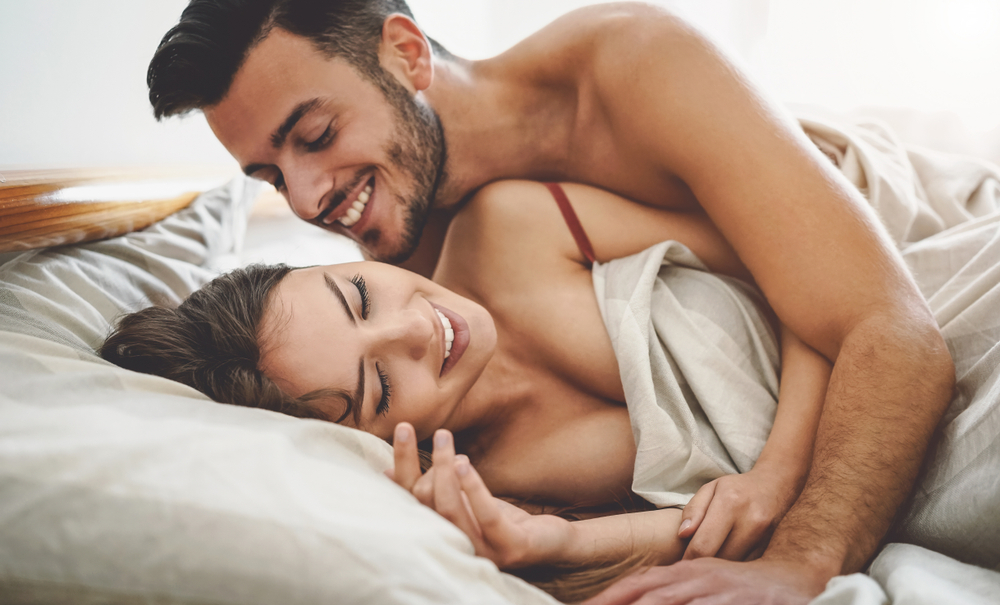 Mann og kvinne i en intim, leken stund i sengen. Hun smiler med lukkede øyne, mens han kysser henne og holder rundt henne med varme.