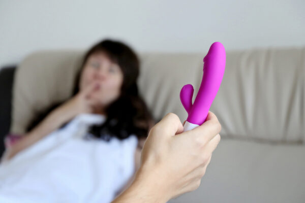 En mann holder en rosa rabbitvibrator mens en kvinne ligger i sengen og smiler forventningsfullt i bakgrunnen.
