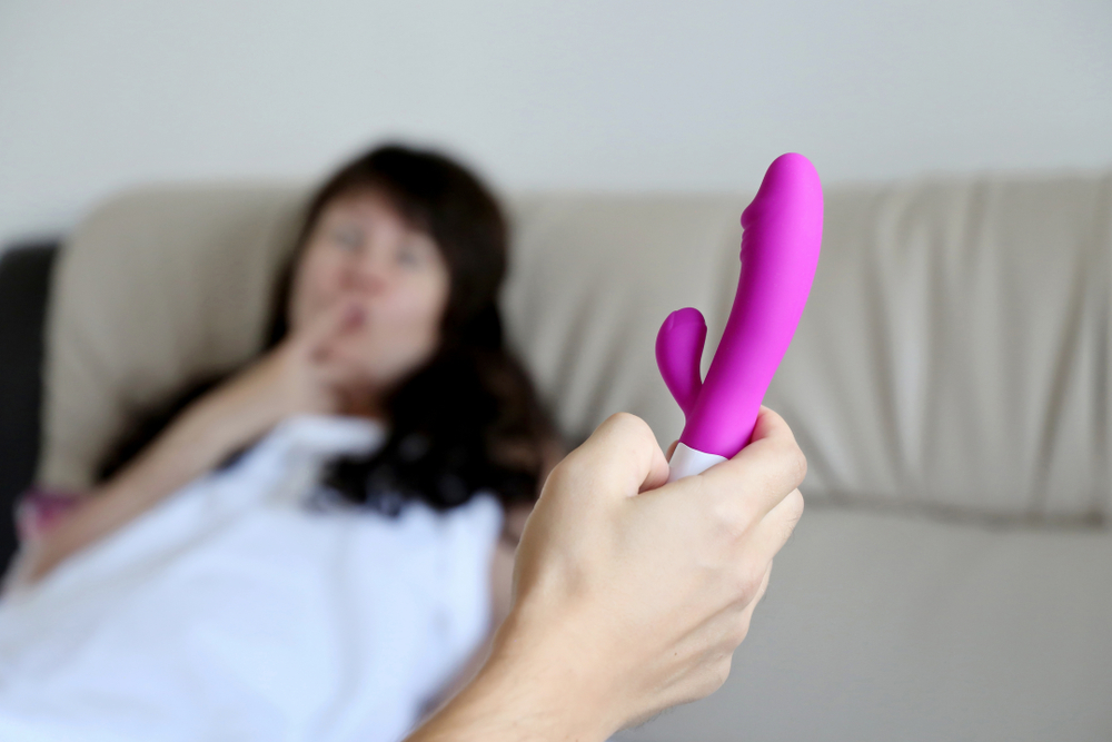 En mann holder en rosa rabbitvibrator mens en kvinne ligger i sengen og smiler forventningsfullt i bakgrunnen.